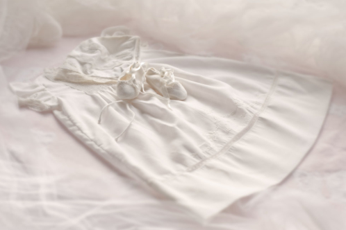 A baby christening gown and booties laid out on a pink silk background