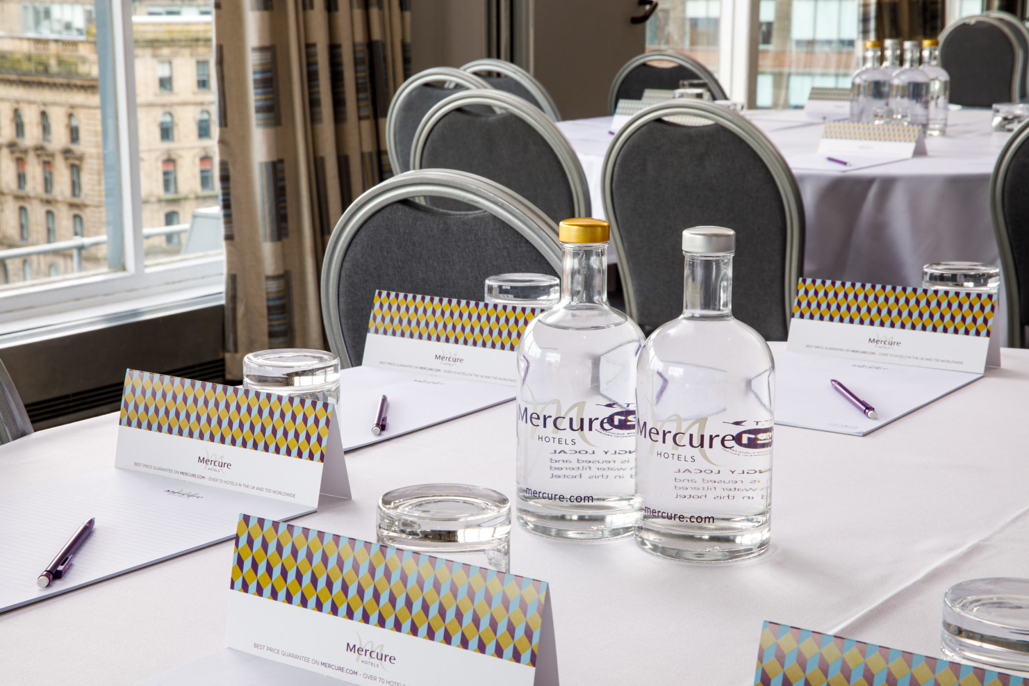 Mercure-Manchester-Piccadilly-Hotel-Park-Suite-Meeting-Room-10-lr-080320 Long table seating 18 in The Congress Room at mercure gloucester bowden hall hotel ready for a meeting, with notepads, bottles of water and bowls of apples on the table